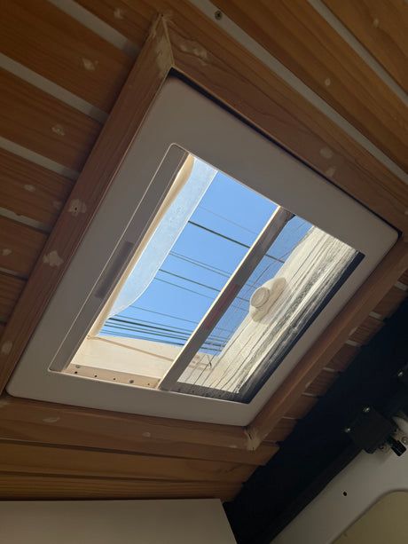 Interior of an rv rental: a wooden ceiling frames an open skylight with an insect screen, revealing bright blue sky and wires.