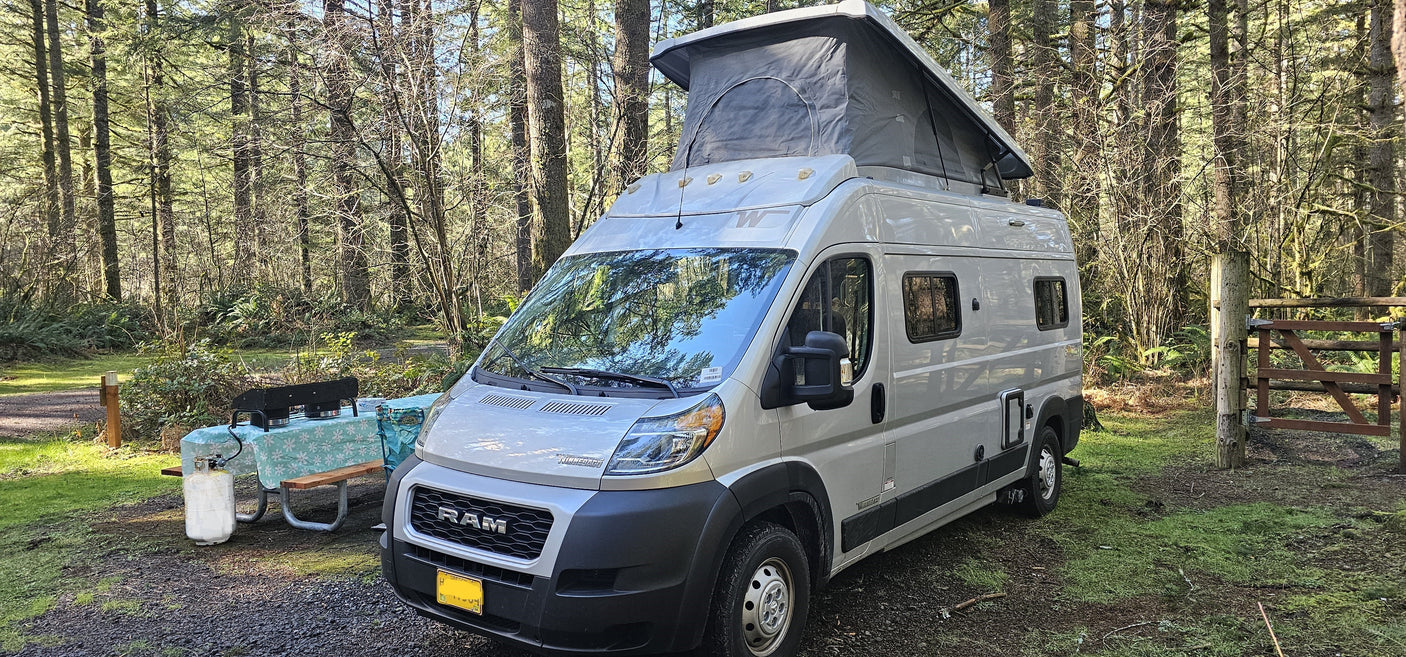 A light grey RV rental camper van with pop-top at a sunlit forest campsite, featuring a picnic table and grill.
