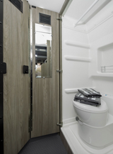Compact RV rental bathroom featuring a toilet with towels, a shower area, and a wooden door with a mirror and vents.