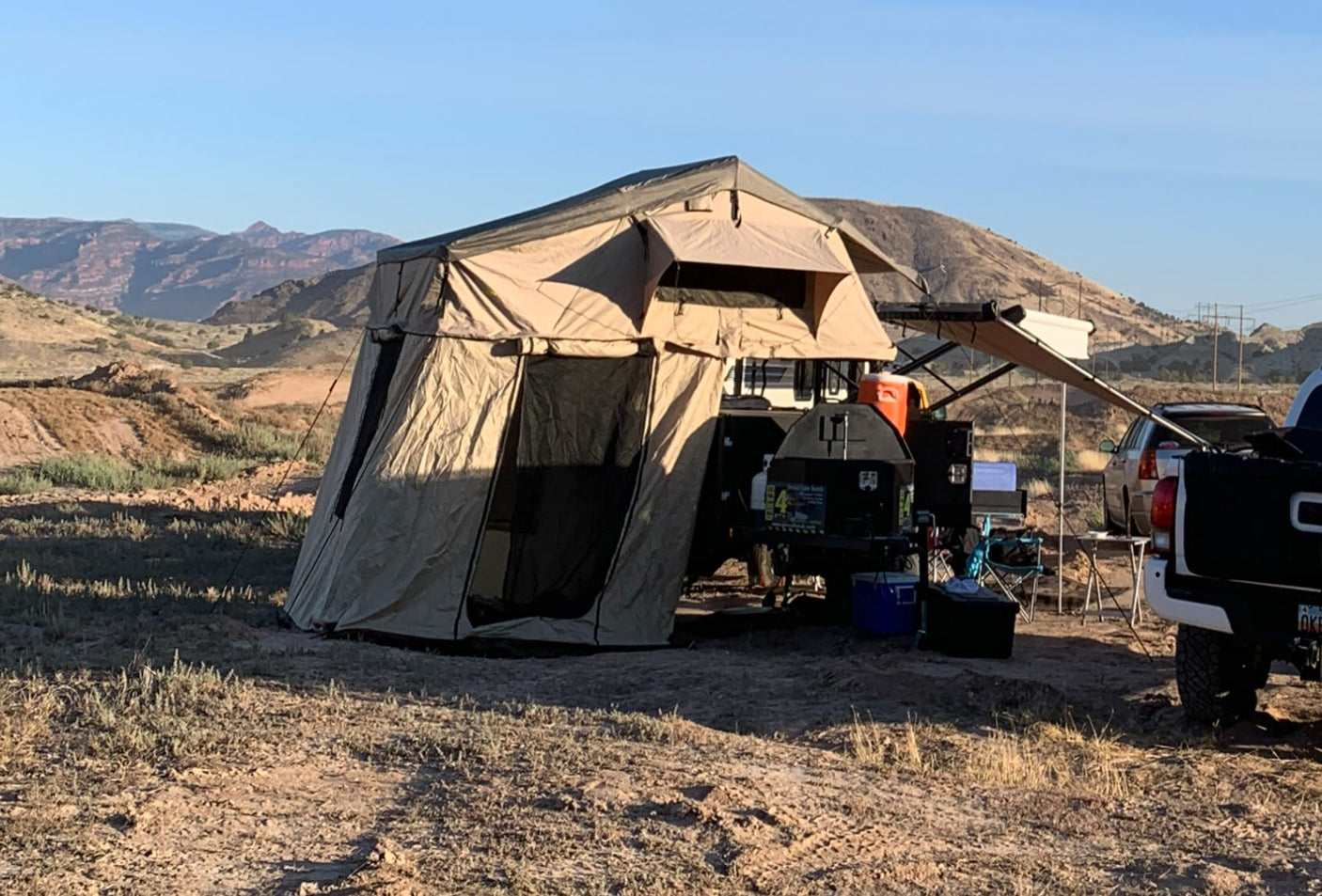 RV rental-style overland camping setup with a rooftop tent, trailer, and truck in a vast, sunny desert landscape.