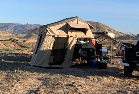 RV rental-style overland camping setup with a rooftop tent, trailer, and truck in a vast, sunny desert landscape.