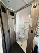 RV rental's compact wet bath: a white toilet inside a shower pan, with a shower curtain and wood-grain walls.