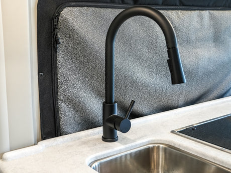 Sleek black faucet & stainless sink in a modern RV rental kitchen, featuring light counters & textured wall panel. Ready for your adventure.