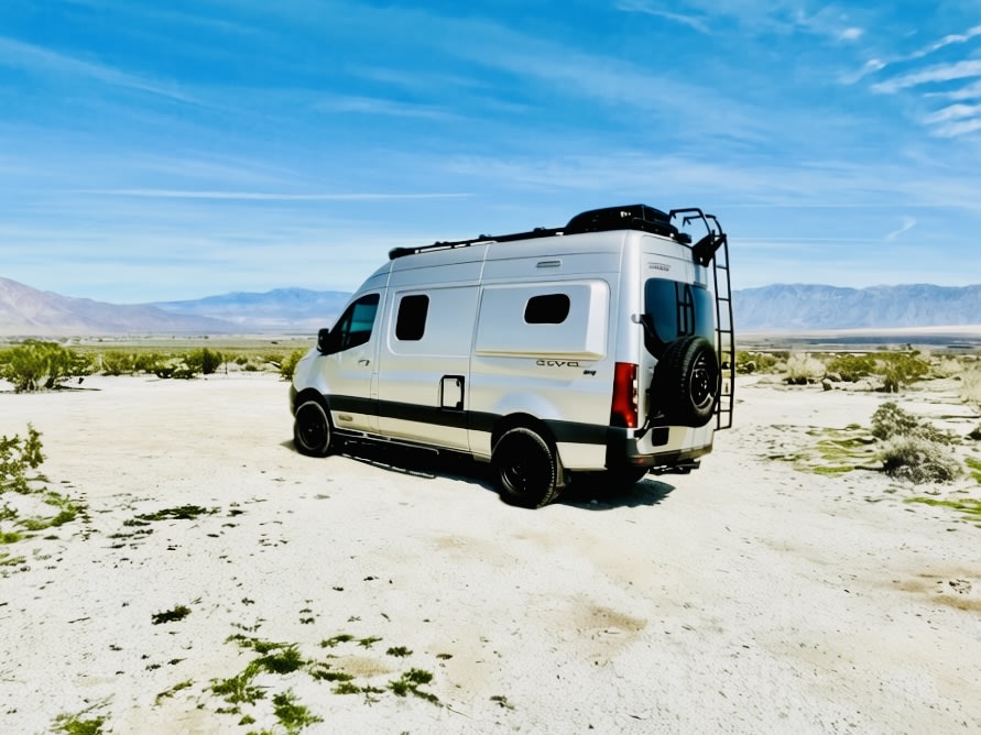 Silver rv rental van in a vast desert with mountains under a blue sky. A perfect rig for off-grid van life adventures.