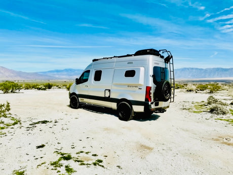 Silver rv rental van in a vast desert with mountains under a blue sky. A perfect rig for off-grid van life adventures.