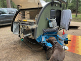 A brown dog rests at a forest campsite, near a teardrop rv rental with its outdoor kitchen open, ready for cooking.