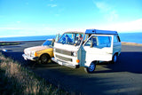 Vintage VW camper van and sedan by the bright blue ocean, great for an RV rental road trip.