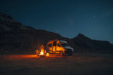 RV rental night camping: A person by a campfire next to a camper van under a vast, star-filled sky, with dark mountains.