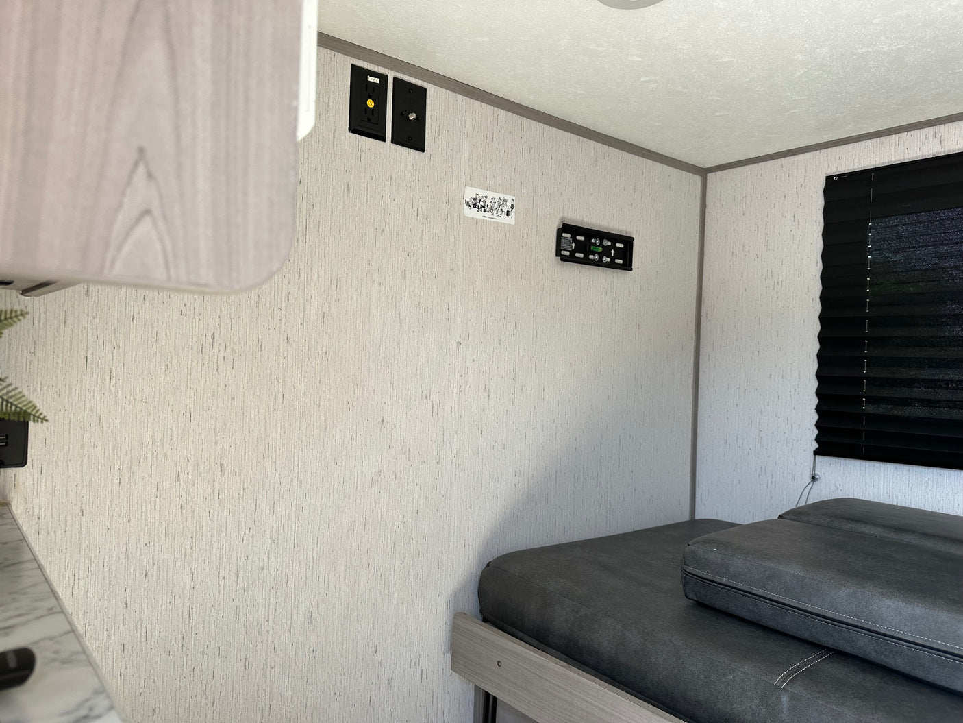 Inside an RV rental, featuring a dark grey bed, TV mount, electrical outlets, and a black window blind.