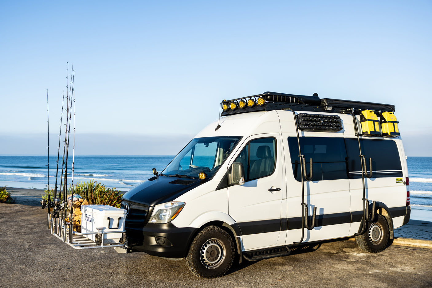 This RV rental Sprinter van, customized for beach adventures, features fishing rods, a cooler, and roof rack gear.