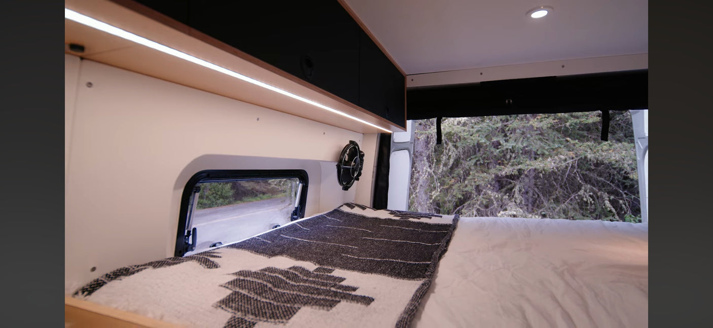 Comfortable RV rental bed with a fan, ambient lighting, and window view of a road and nature in a camper van.