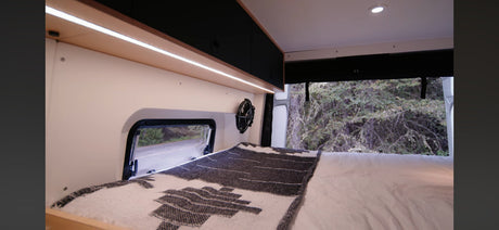 Comfortable RV rental bed with a fan, ambient lighting, and window view of a road and nature in a camper van.