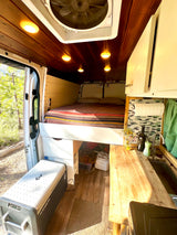 RV rental interior with a cozy bed, compact kitchen, smart storage, and natural light from the open side door.