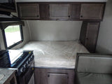 Compact RV rental interior: features an unused bed in plastic, kitchen stove/oven, sink area, and overhead storage.