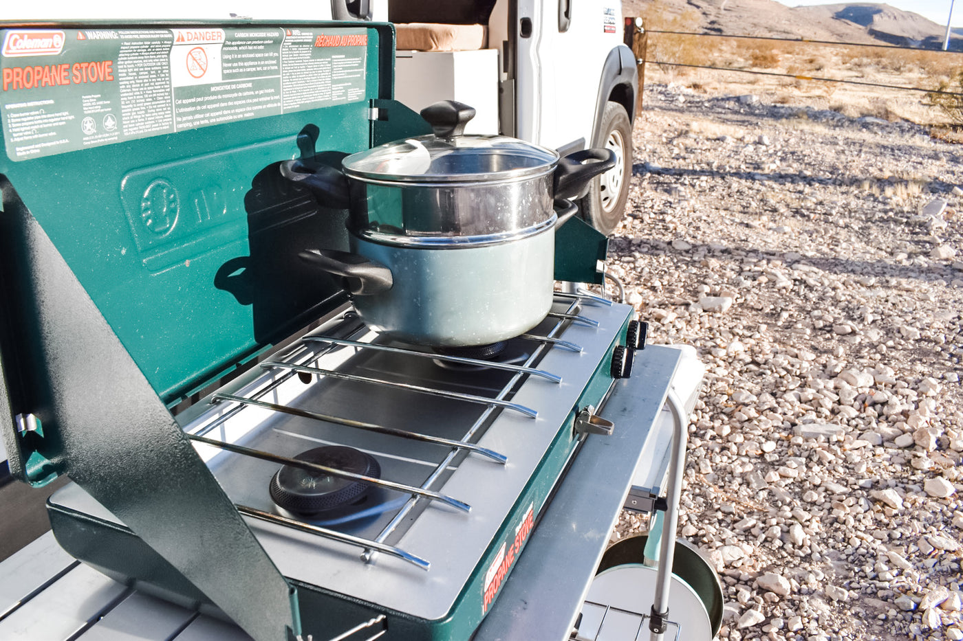 RV rental trip: Cooking outdoors on a Coleman propane stove with a pot, set up next to a camper van on gravel. — photo 4