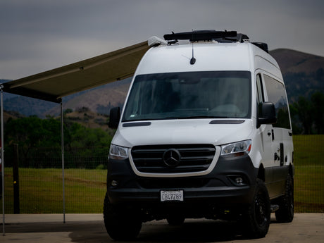 This white Mercedes Sprinter van RV rental, with an extended awning, is parked outdoors against a scenic, hilly backdrop.
