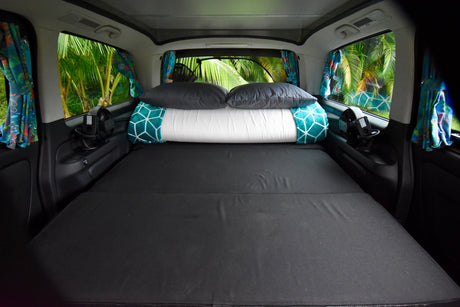 This RV rental campervan's cozy bed setup: black mattress, pillows, fans, and vibrant tropical views from the windows. Ready for adventures.