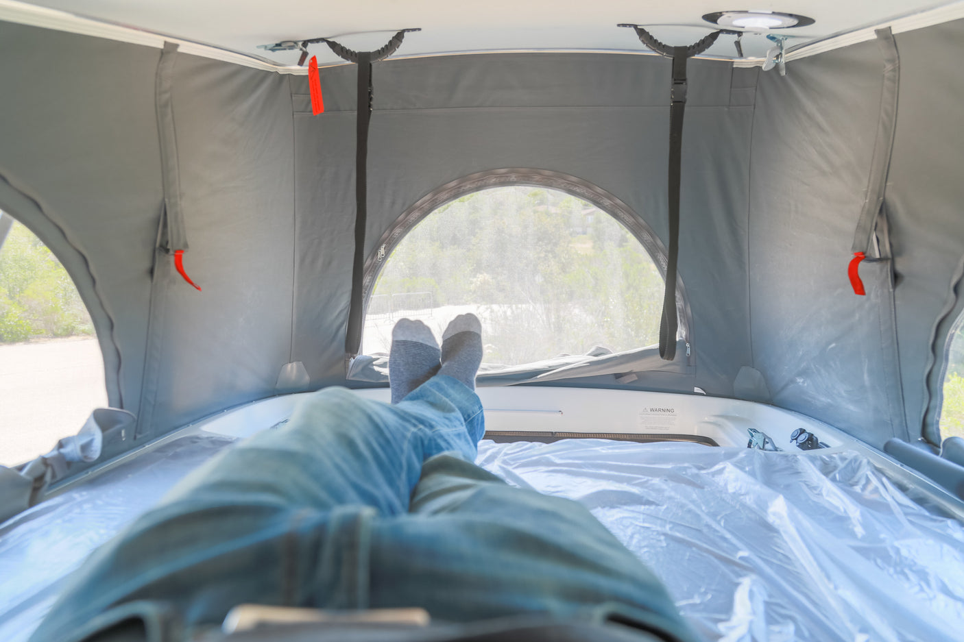 Person relaxing, feet up, inside a pop-top RV rental, comfy with grey canvas walls & a mesh window view.