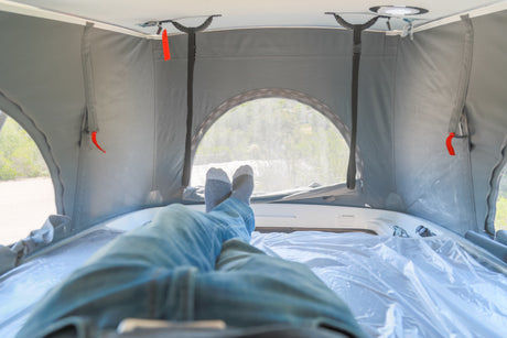 Person relaxing, feet up, inside a pop-top RV rental, comfy with grey canvas walls & a mesh window view.