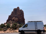 A white Sprinter-style **rv rental** van with a roof rack and ladder parked next to large red rock formations in a sun-drenched desert.