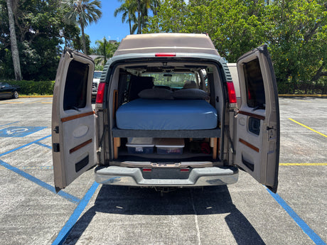 Rear view of an RV rental conversion van with open doors, showing a comfortable bed and storage inside. Ideal for travel.