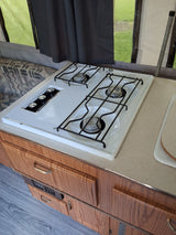 An RV rental kitchen showcasing a clean, white three-burner gas stovetop, wood cabinetry, and a bright window view.