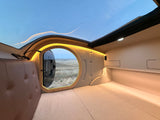 Cozy RV rental interior with a panoramic roof and a unique porthole window overlooking scenic fields.