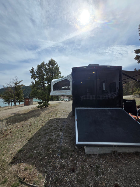 Scenic rv rental at a lakeside campsite, featuring a toy hauler with open ramp and pop-out tent. Bright sun overhead.