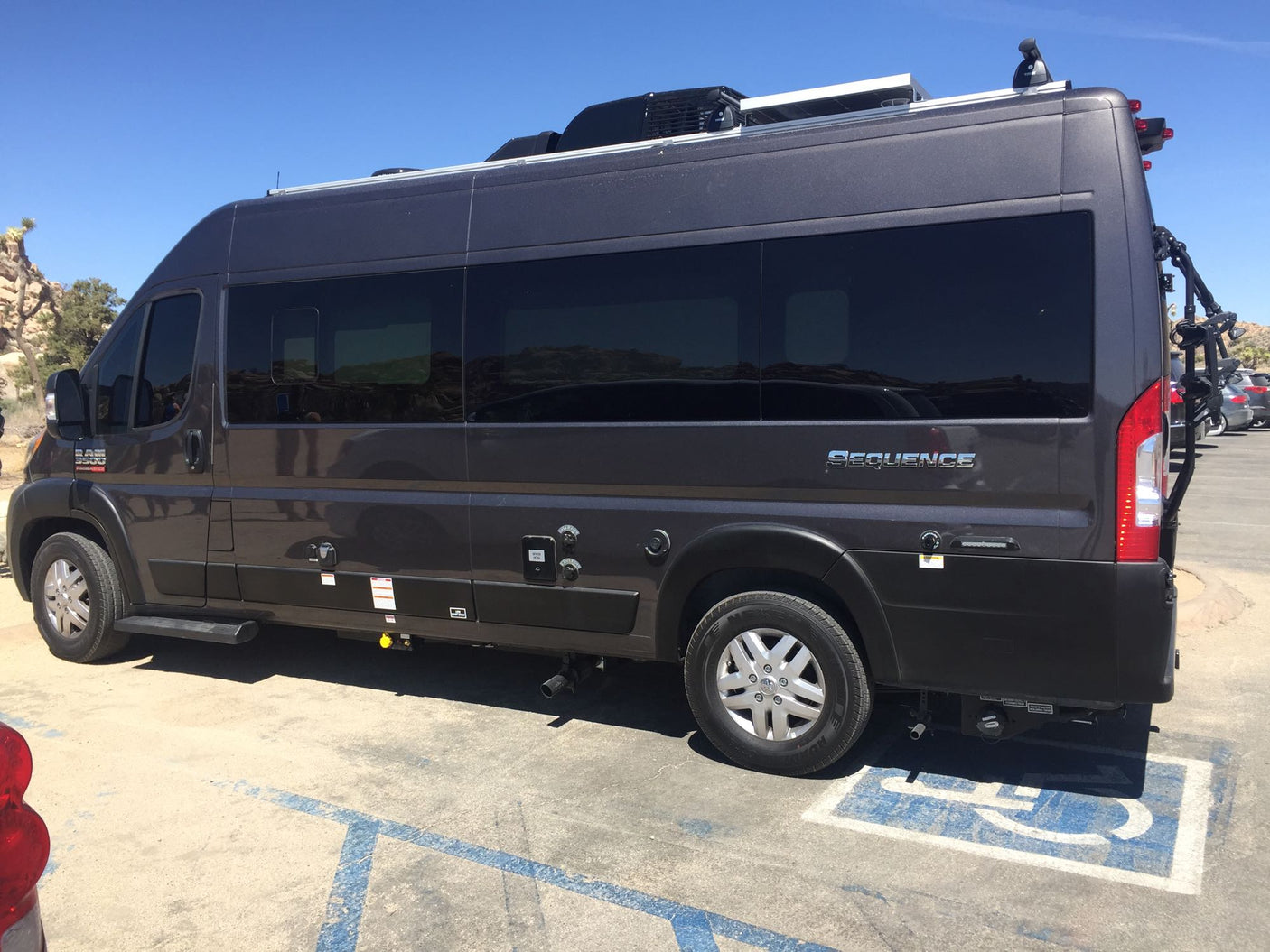 Dark gray Ram ProMaster camper van, an RV rental, parked in a handicapped spot with rooftop solar & bike rack.