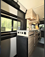 This RV rental interior features a compact kitchen with a sink, refrigerator, wood cabinets, and large windows overlooking trees.