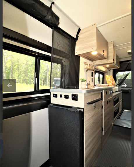 This RV rental interior features a compact kitchen with a sink, refrigerator, wood cabinets, and large windows overlooking trees.