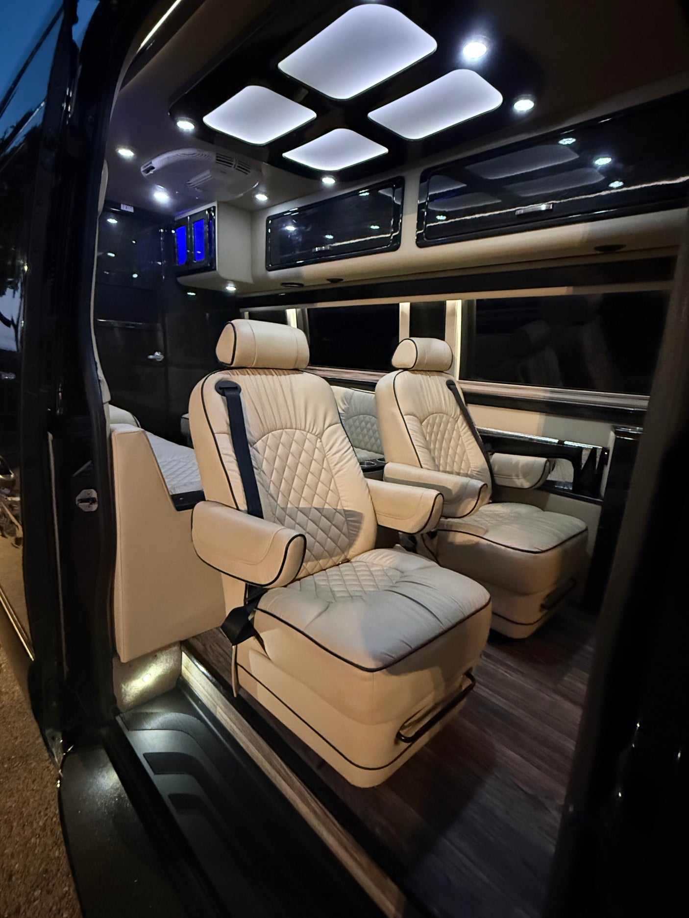 Step inside a premium RV rental: spacious custom van interior with cream captain's chairs, wood flooring & LED lighting.
