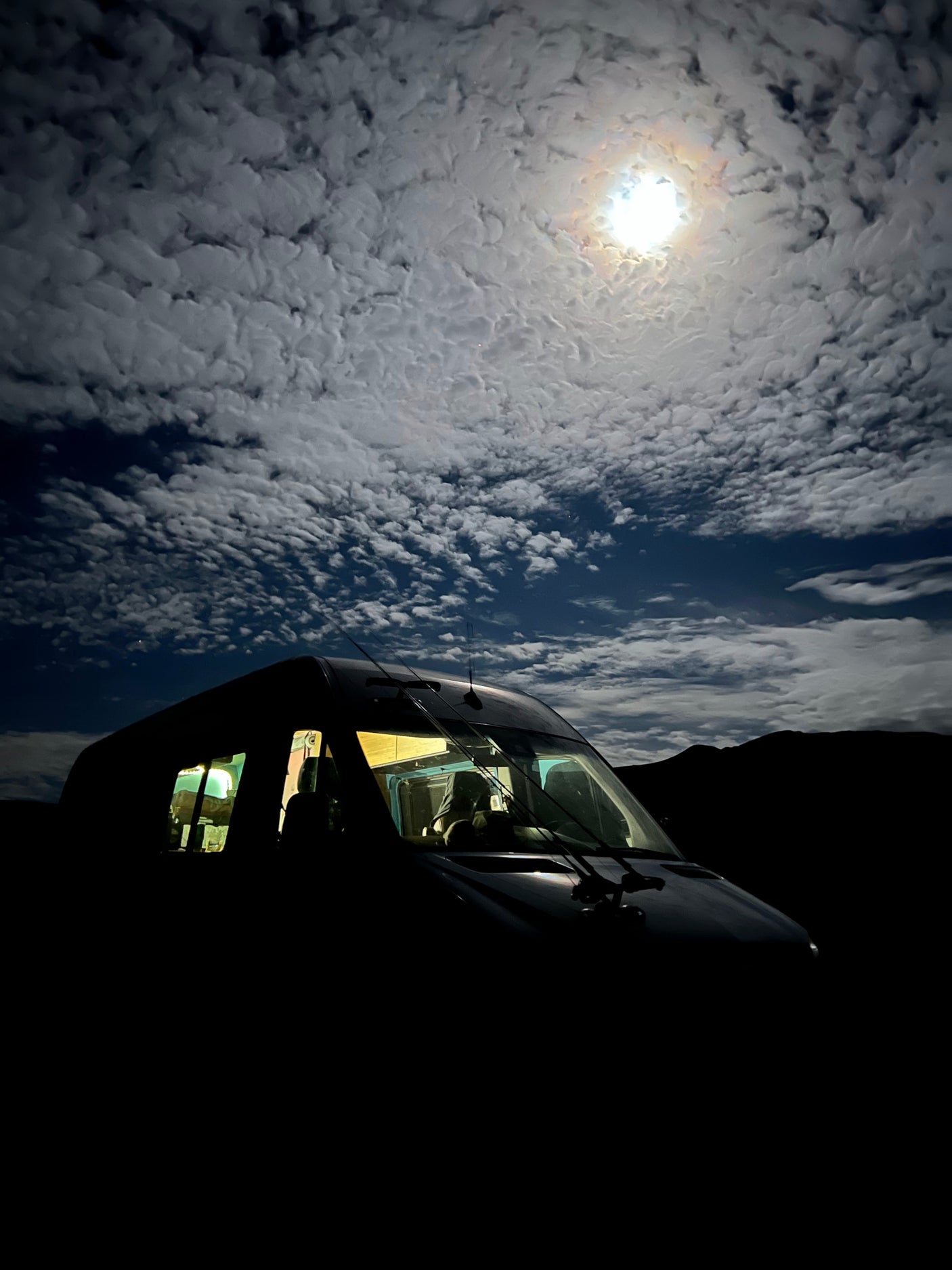 Experience moonlit nights in an RV rental! This dark camper van glows inside, parked under a brilliant moon & clouds, mountains silhouetted.