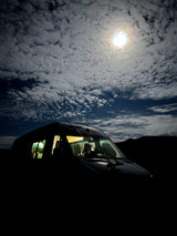 Experience moonlit nights in an RV rental! This dark camper van glows inside, parked under a brilliant moon & clouds, mountains silhouetted.