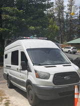 This white Ford Transit camper van, an ideal RV rental, is parked outdoors among tall trees, ready for adventure.