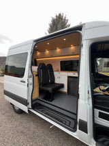 Step inside this modern white camper van's custom interior, featuring seating, kitchen, and bed via its open side door. Perfect for rv rental!