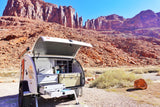 Open RV rental teardrop trailer showing its outdoor kitchen with a Yeti cooler, parked in a scenic red rock desert campsite.