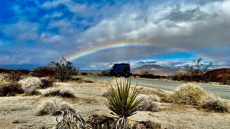 Experience adventure! An RV rental van on a desert road with a vibrant rainbow overhead, mountains, and desert plants.