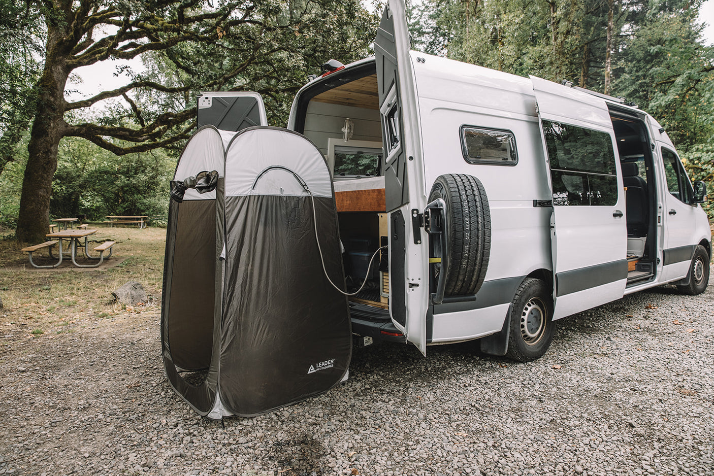 An RV rental van with open rear doors reveals its interior, while an outdoor privacy tent provides a shower at a scenic campsite.