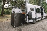 An RV rental van with open rear doors reveals its interior, while an outdoor privacy tent provides a shower at a scenic campsite.