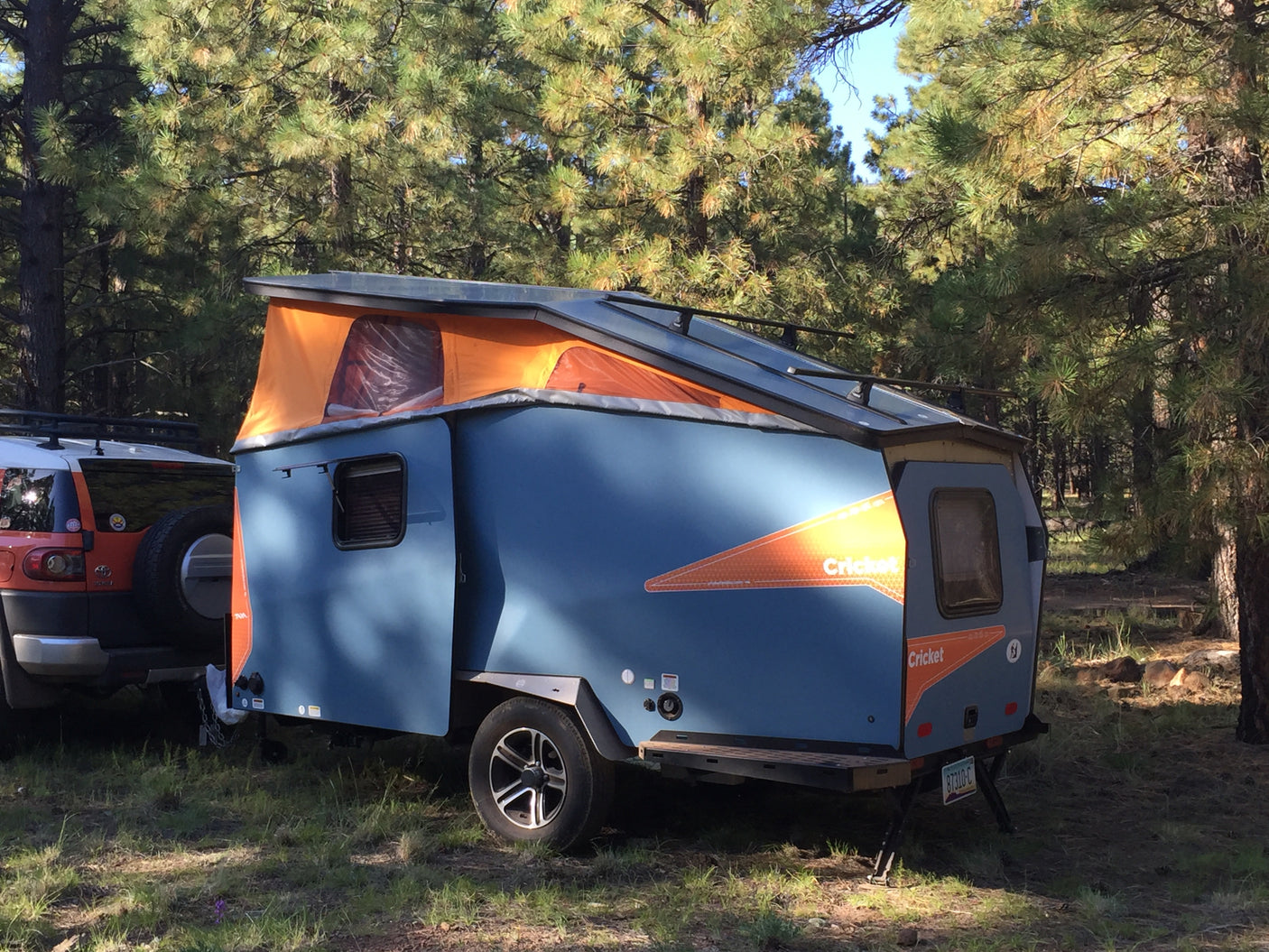 A blue Cricket pop-up camper attached to an SUV in a sunny pine forest, perfect for an rv rental adventure.