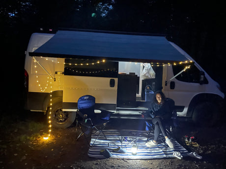A person relaxes by their white RV rental camper van at night. String lights illuminate the awning, creating a cozy outdoor camping scene.