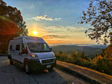 Scenic mountain view with a white RV rental parked on asphalt, bathed in golden sunrise light over misty valleys.