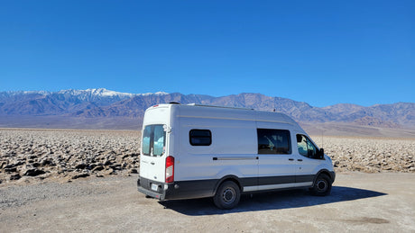 White RV rental parked at a unique, rocky desert landscape. Snow-capped mountains under a clear blue sky.