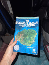 Ready for an RV rental trip to Kauai? A hand holds "The Ultimate Kauai Guidebook" inside a vehicle.