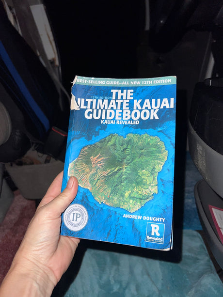 Ready for an RV rental trip to Kauai? A hand holds "The Ultimate Kauai Guidebook" inside a vehicle.