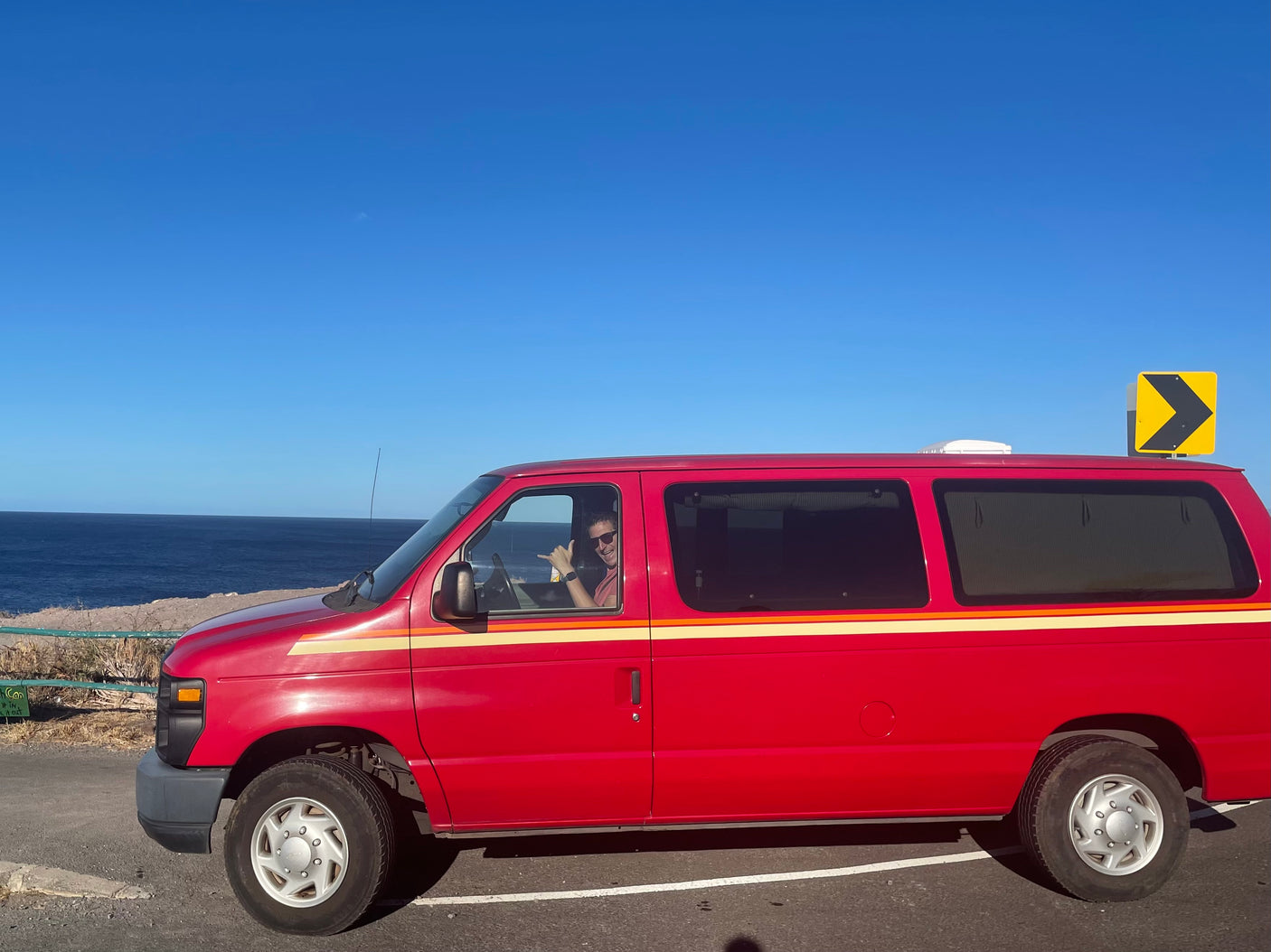 A red RV rental van is parked at a scenic ocean overlook under a clear blue sky. The driver smiles, making a shaka sign.