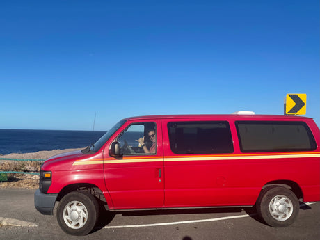 A red RV rental van is parked at a scenic ocean overlook under a clear blue sky. The driver smiles, making a shaka sign.