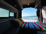 RV rental: Comfy bed with colorful serape blanket & pillows, facing ocean waves on a sandy beach. Ultimate van camping.