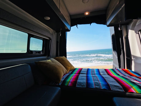 RV rental: Comfy bed with colorful serape blanket & pillows, facing ocean waves on a sandy beach. Ultimate van camping.